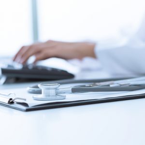 A doctor's hands on a keyboard in the background with a stethoscope and clipboard in the foreground.