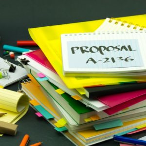 A stack of red and yellow files and notebooks. On the top notebook, "Proposal A-2136" is written in black marker, referencing Michigan's proposed Medicaid work requirements.