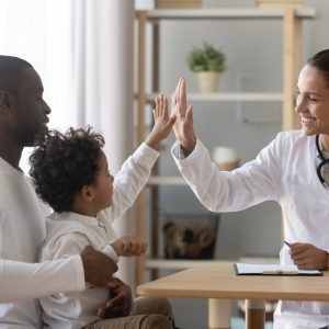 A physician high-fives a child who is sitting on his father's lap. The father is an example of the Michigan residents the 2018 Cover Michigan Survey speaks with.