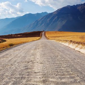 Image of a rural road with fields on both side and mountains in the background.