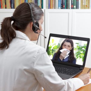 A doctor sits in front of a laptop, offering telehealth services during COVID-19. The patient shown in the laptop holds a tissue to her nose..