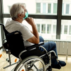 An older adult with a disability looks out a window, seeming lonely and wearing a mask.