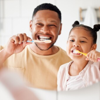 A father and daughter brush their teeth, smiling.