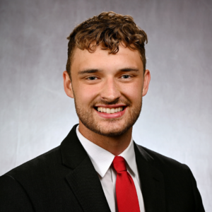 Adam has short brown hair, curly and longer in the front. He is wearing a black blazer, white button up shirt, and red tie. He is smiling.