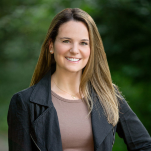 Andrea has light brown hair with highlights. She is wearing a blue shirt and black jacket. She is smiling with one hand on her hip. There is a tree in the background.