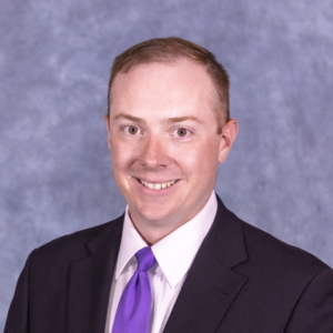 Connor has closely cropped brown hair. He is wearing a black jacket, white button up shirt, and bright purple tie. He is smiling against a light purple backdrop.