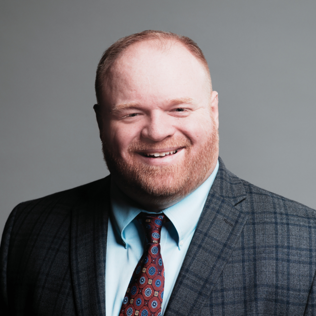 Daniel has reddish brown hair and beard. He is wearing a dark blue plaid blazer, light blue button up shirt, and burgundy necktie. He is smiling against a grey background.