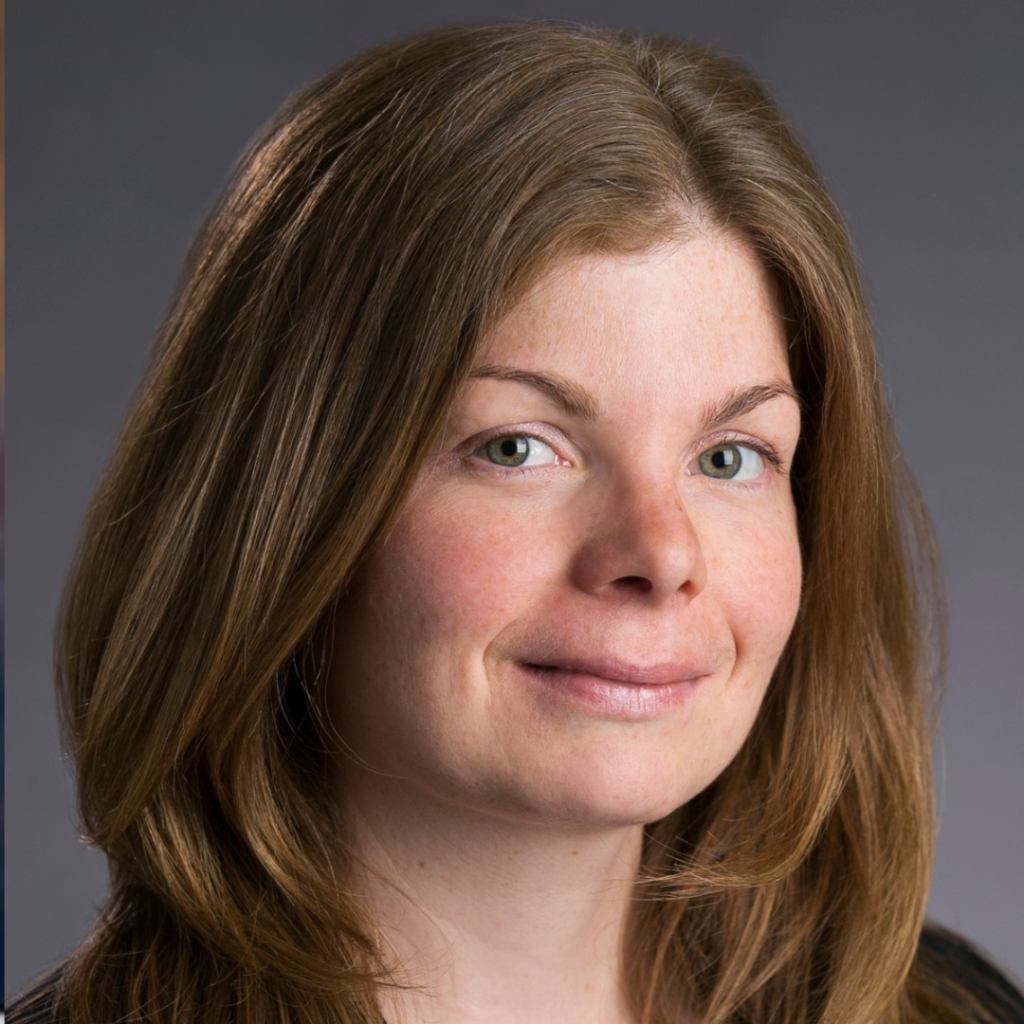 Julie has reddish brown hair parted on the side. She is smiling against a grey backdrop.