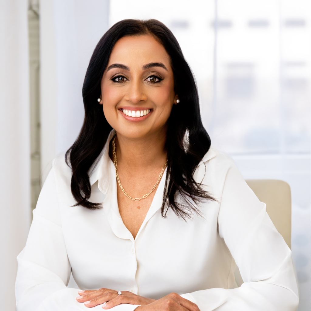 Minal has dark hair and a white button up shirt. She is smiling with her hands crossed on the table in front of her. There is a building outside the window in the background.