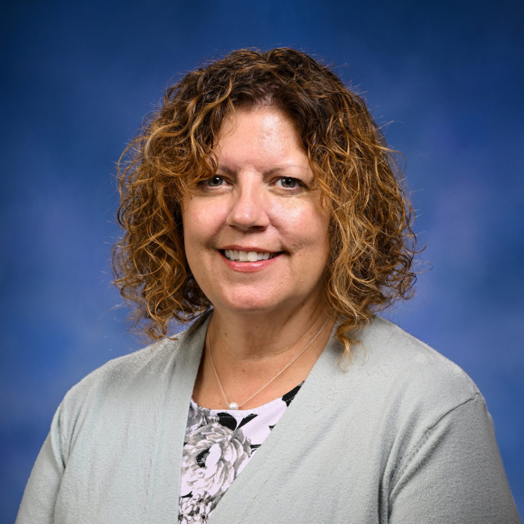 Mindy wears a pale gray sweater and pink shirt. She is smiling against a blue backdrop.