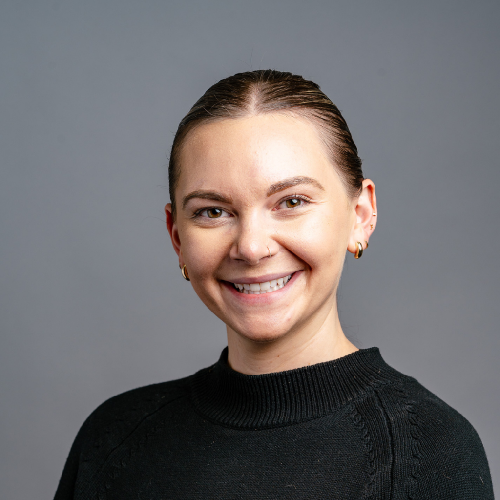 Stephanie's brown hair is parted in the middle and pulled back in a ponytail or bun. She wears a black shirt and is smiling against a grey backdrop.