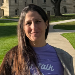 Taryn has long brown hair and wears a purple t-shirt. She is smiling with a historic building in the background.