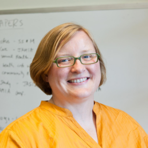 Tiffany has reddish blond hair and glasses. She is wearing a bright yellow shirt and glasses. She is smiling. In the backround is a whiteboard with writing on it.